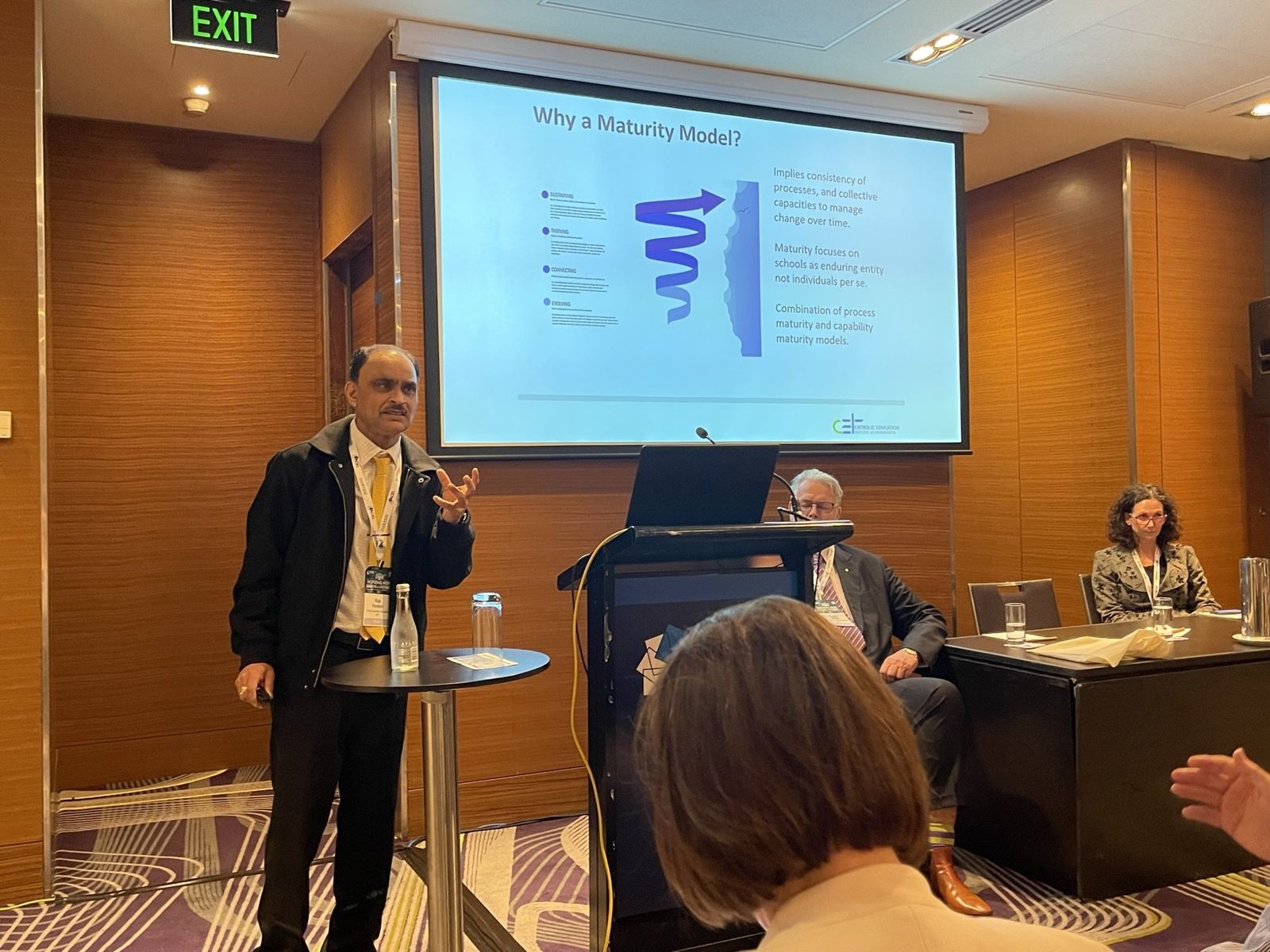 Dr. Raju Varanasi speaking beside a slide titled Why a Maturity Model